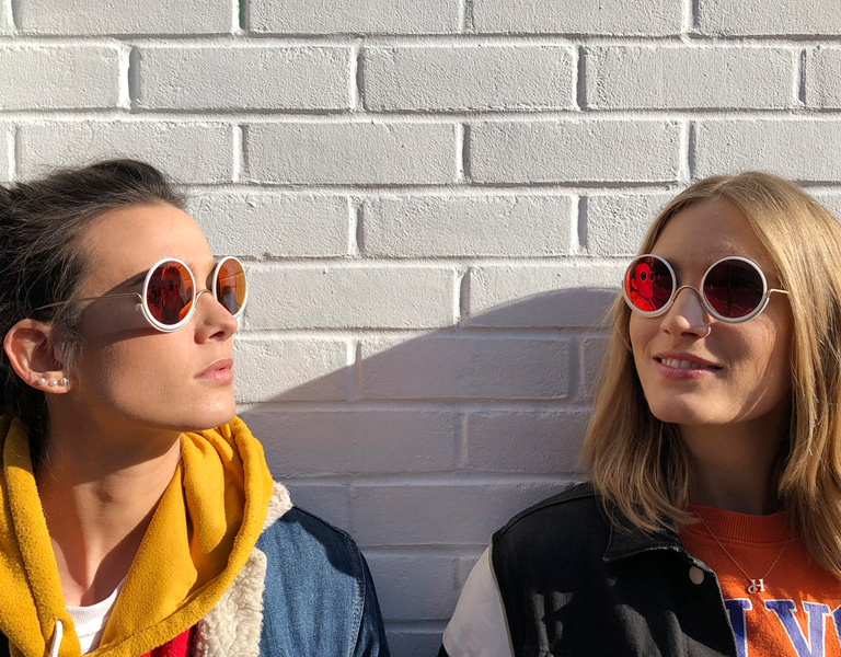 image of two women wearing Wire Glasses sunglasses leaning against a white brick wall